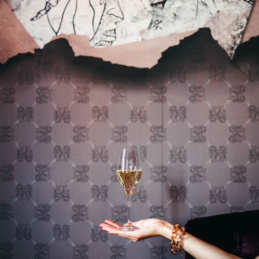 En hand som balanserar ett vinglas med vitt vin i. På armen finns ett guld armband