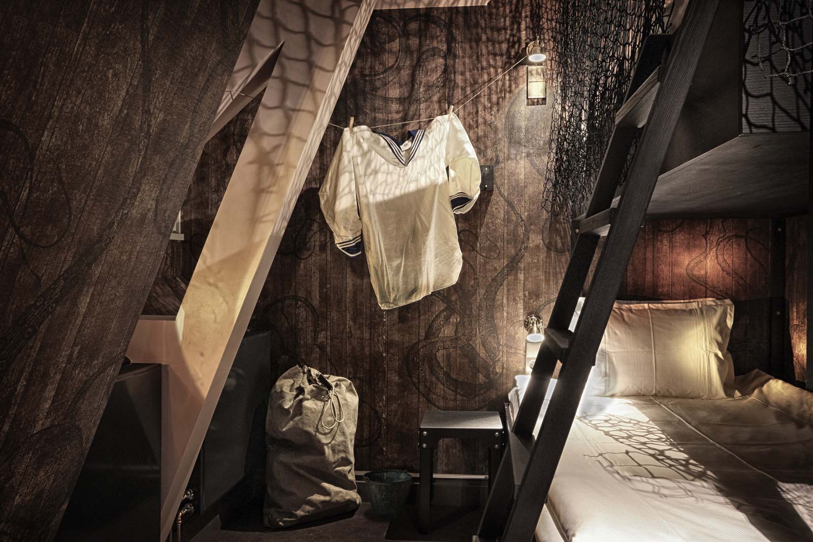 Kosy hörn i hotellrum med upphängd vit skjorta, en stege som leder till en loft-säng under ett fönster som släpper in mjukt ljus.