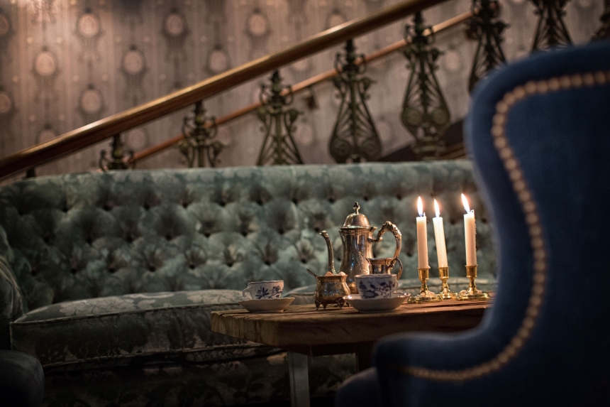 Del av en hotellounge med en djupblå sammetsfåtölj och en tuftad soffa, accentuerad av ett kaffebord med tekanna och tända ljus som framhäver en avkopplande atmosfär.