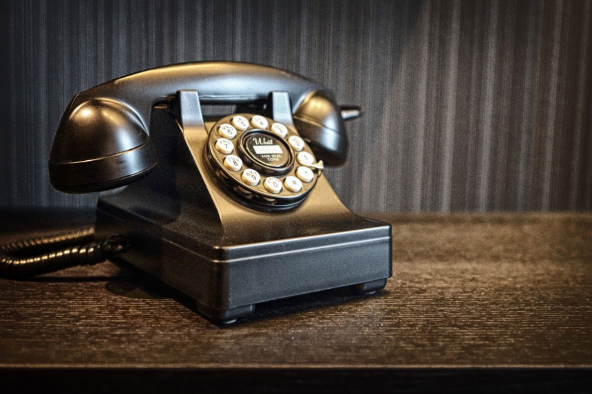 Vintage svart telefon med vridskiva på ett mörkt träbord framför en grå texturerad bakgrund.